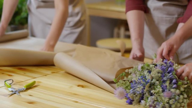 Pan close-up hands shot of three female florists at work in flower shop, one spreading packaging paper on wooden table, her colleagues wrapping freshly made wildflower bouquet and tying it with twine