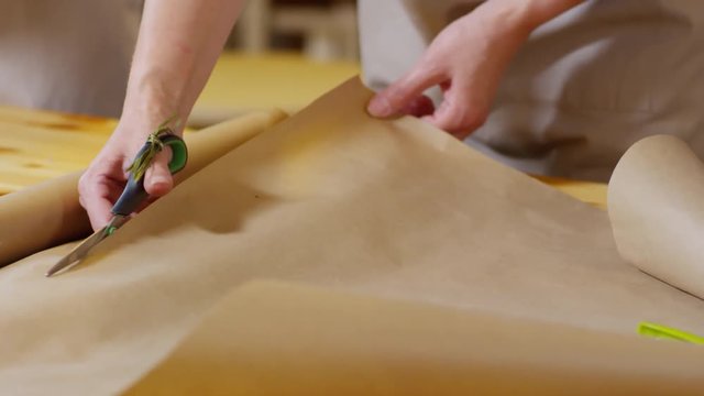 Close-up hands shot of lady florist with natural manicure, wearing beige apron, carefully cutting plain brown wrapping paper with scissors on wooden worktop