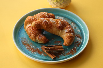 Two croissants in a blue plate on a yellow background. Cocoa crumb and flour sprinkled next and a little cinnamon
