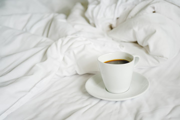 white ceramic cup of hot black coffee on messy bed sheet, in soft focus