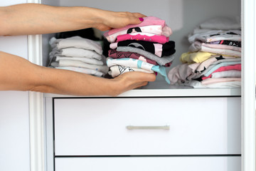 Mom picking out kid outfit at home to send her children back to school. New colored clothing stacked in a pile close-up in white wardrobe. Woman folding clothes on a shelf in a closet.
