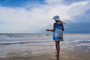 A girl in a beautiful dress and a white hat with a shell in her hands on the shore against the blue sky background
