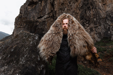 a serious and strict man with a beard and mustache in a black T-shirt and a bear skin stands at a cliff, a mountain.