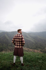 man kilt with mustache beard standing on the top Green Mountain grass, dressed plaid shirt, white socks brown shoes, looks around. it is snowing from the sky. fog around.Caucasian