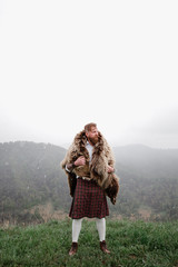 man with mustache beard is standing on top mountain kilt. bear's skin on the back. in a white shirt, socks a tattoo on his arm. on the grass. there is snow.leather bracelet