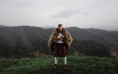 man with mustache beard is standing on top mountain kilt. bear's skin on the back. in a white shirt, socks a tattoo on his arm. on the grass. there is snow.leather bracelet - obrazy, fototapety, plakaty