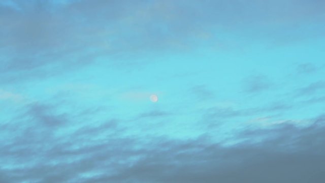 Zooming out on a small moon in the sky surrounded by clouds, taken early evening