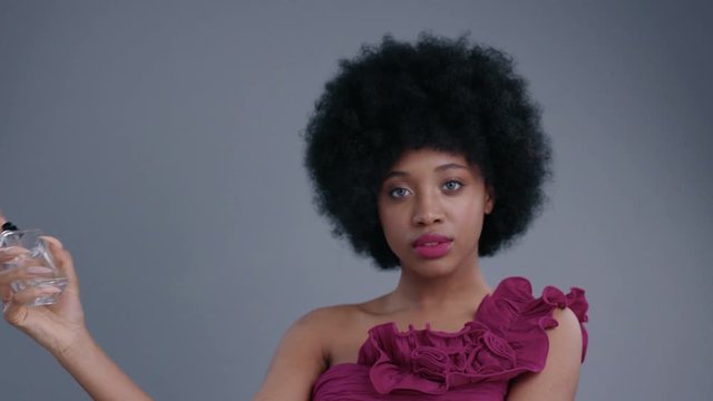 Elegant female model with pink lips and dress spraying perfume isolated on grey background. Portrait of beautiful afro-american girl promoting nice women's fragrance.