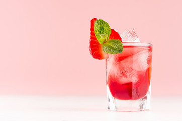 Cold red alcoholic liquor with ice cubes, strawberry slice, green mint in shot glass on white wood...