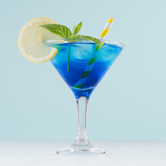 Blue curacao cocktail with ice cubes, lemon slice, yellow straw, green mint in luxury martini glass on white wood board and pastel mint color wall, square.