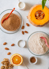 Ingredients for baking healthy cake: pumpkin, whole wheat flour, brown cane sugar, walnuts and almonds, vegan milk and orange. Top view, the layout on a light concrete surface.