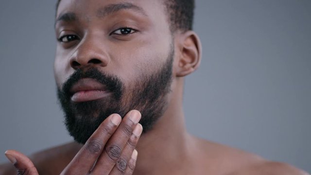 Portrait of confident sexy young black man touching his beard looking in the mirror. Close-up of handsome shirtless guy readyto shave in his bathroom at home.