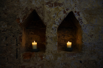 Fototapeta na wymiar two white burning candles in two niches in a dark old brick wall of a monastery, copy space