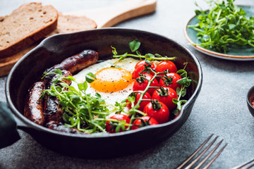 Breakfast time, fried egg with sausages and cherry tomatoes in a black iron pan, served microgreens.