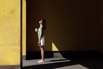 Attractive young woman with take-away drink standing against yellow wall