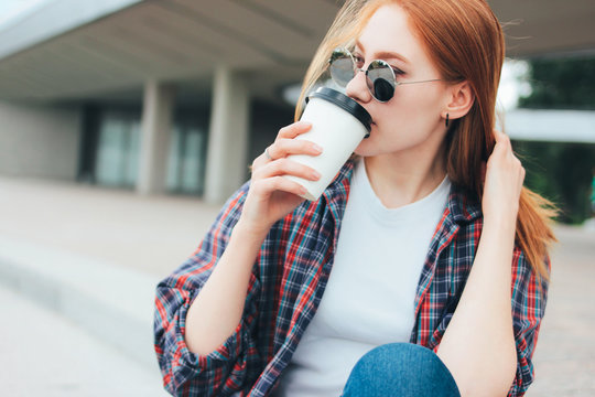 Attractive redhead smiling girl in round sunglasses casual wearing drinking coffee to go sitting on street in city