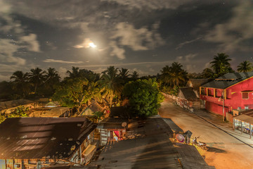 Manakara, Madagascar street view at night. Dirt road street in between houses and tropical vegetation with moonlight, stars and clouds in the sky