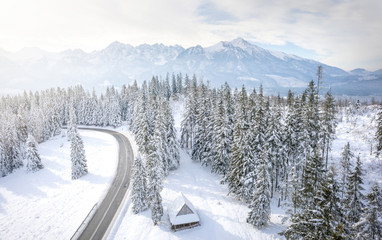 Snowy winter mountains landscape with frosty forest and road. Road to rocky mountain range. Wooden house in pine trees in mountain village