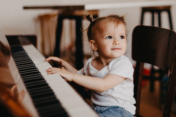 a portrait of a cute 2-year-old happy baby who has fun at the piano lesson. Little child girl playing piano in a light room. Selective focus, noise effect