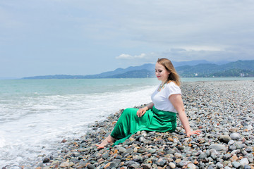 Amazing woman sitting on stones, on the beach near sea. Sincere emotions from sea waves. Plus size woman is dressed in a long green skirt and a white blouse. And a big colored necklace on the neck.