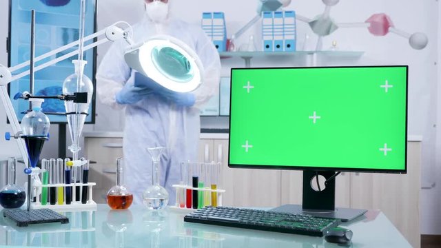 Computer with green screen mock-up in secure research facility. A chemist is walking in the background