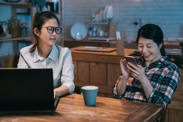 Angry business worker woman looking at lazy mate playing with the smart phone. annoyed young girl roommate friend laughing loudly sitting next to hard working employee in home kitchen. mad lady.