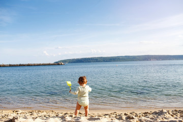 A small child stands on the beach with a toy in his hands. The baby will soon be swimming in the sea.