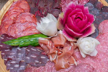 sliced assorted cured meat and sausages decorated with carved flowers of vegetables on a wooden plate
