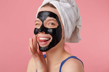 Portrait of young happy woman after shower with a towel on her head, with black mask, touches face and smiles, winks and looks at the camera stands over pink background.