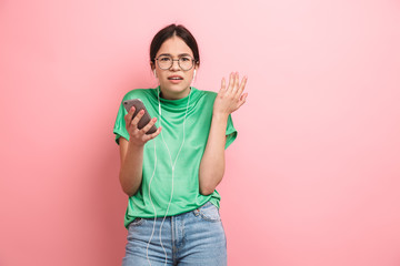 Photo of displeased young girl wearing round eyeglasses using earphones while holding smartphone