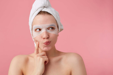 Close up of thinking young woman with a towel on her head after shower, looks away and touches cheek, can't make a decision, stands over pink background.