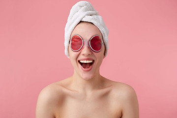 Wow! So cool! Young happy amazed woman after spa mask on eyes and with a towel on her head, with wide open mouth and eyes, hears cool news, stands over pink background.