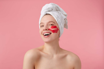 Photo of cheerful young woman after spa with a towel on her head, with patch for lips on cheeks, broadly smiles, feels so happy, stands over pink background.