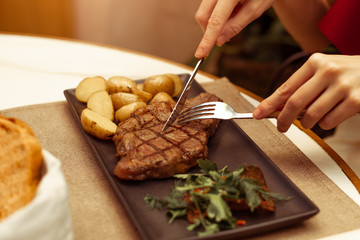 Focus on the meat where the fork and knife. Beautiful female hands in a restaurant at the table cut beef striploin steak.