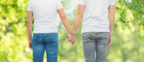 lgbt, same-sex relationships and homosexual concept - close up of male couple wearing gay pride rainbow awareness wristbands holding hands over green natural background