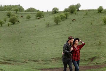 a man with a beard and mustache in a leather jacket and a woman in a plaid shirt in hats are holding hands, on green hills, a ranch. horses are walking.caucasian