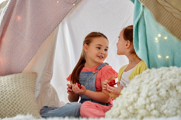 childhood and hygge concept - happy little girls playing tea party with toy crockery in kids tent at home