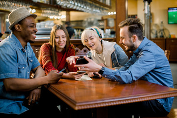 Portrait of cheerful young friends looking at smart phone while sitting in cafe. Mixed race people sitting at a table in restaurant using mobile phone.