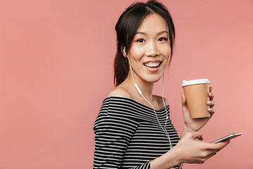 Image of pretty asian woman dressed in basic wear holding paper cup and mobile phone while listening to music with earphones