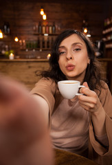 Cheerful beautiful woman giving kissing and taking a self-portrait with smartphone in a coffee shop