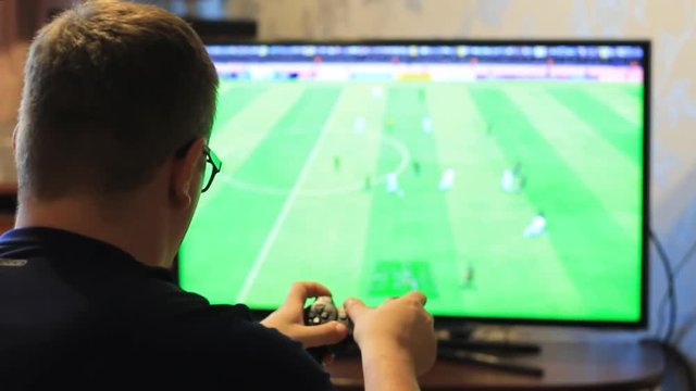 The guy holds a game joystick in his hands and plays football. Blurred monitor screen.