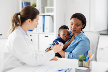 medicine, healthcare and pediatry concept - african american mother with sick baby son and caucasian doctor with thermometer at clinic