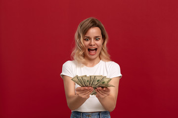 Portrait of a girl with curly blond hair dressed in a white t-shirt standing on a red background. Happy model looks at the bundle of dollars. Excited girl expresses emotion of surprise