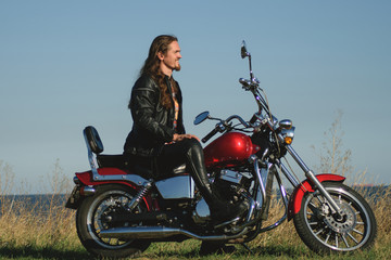 Fototapeta na wymiar Handsome long-haired biker in the leather on red motorcycle against field and seashore. Brutal man in leather