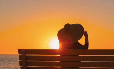 woman in a big hat sits on a bench and looks at the sunset