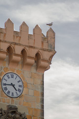 Fototapeta na wymiar Seagull resting on top of a stone tower with a clock and a cloudy sky in the background