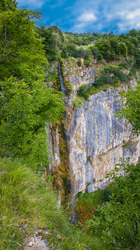 A small waterfall on a rocky leap with green trees and bushes under blue sky with smooth clouds
