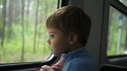 The boy is traveling by train and looks out the window, watching the moving objects outside the window. Travelling with family