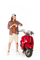Fototapeta na wymiar full length view of young man looking and pointing with finger at watch while standing near red scooter isolated on white