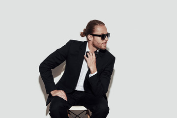 Always perfect. Charming young man in full suit looking away and adjusting bow tie while sitting against grey background